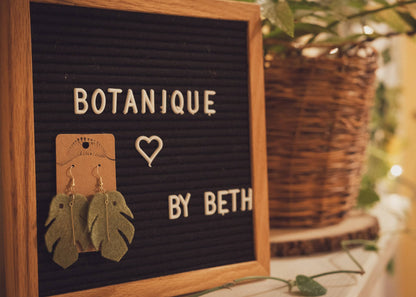 Jade Monstera Leaf Felt Earrings hanging on a sign with plants in the background