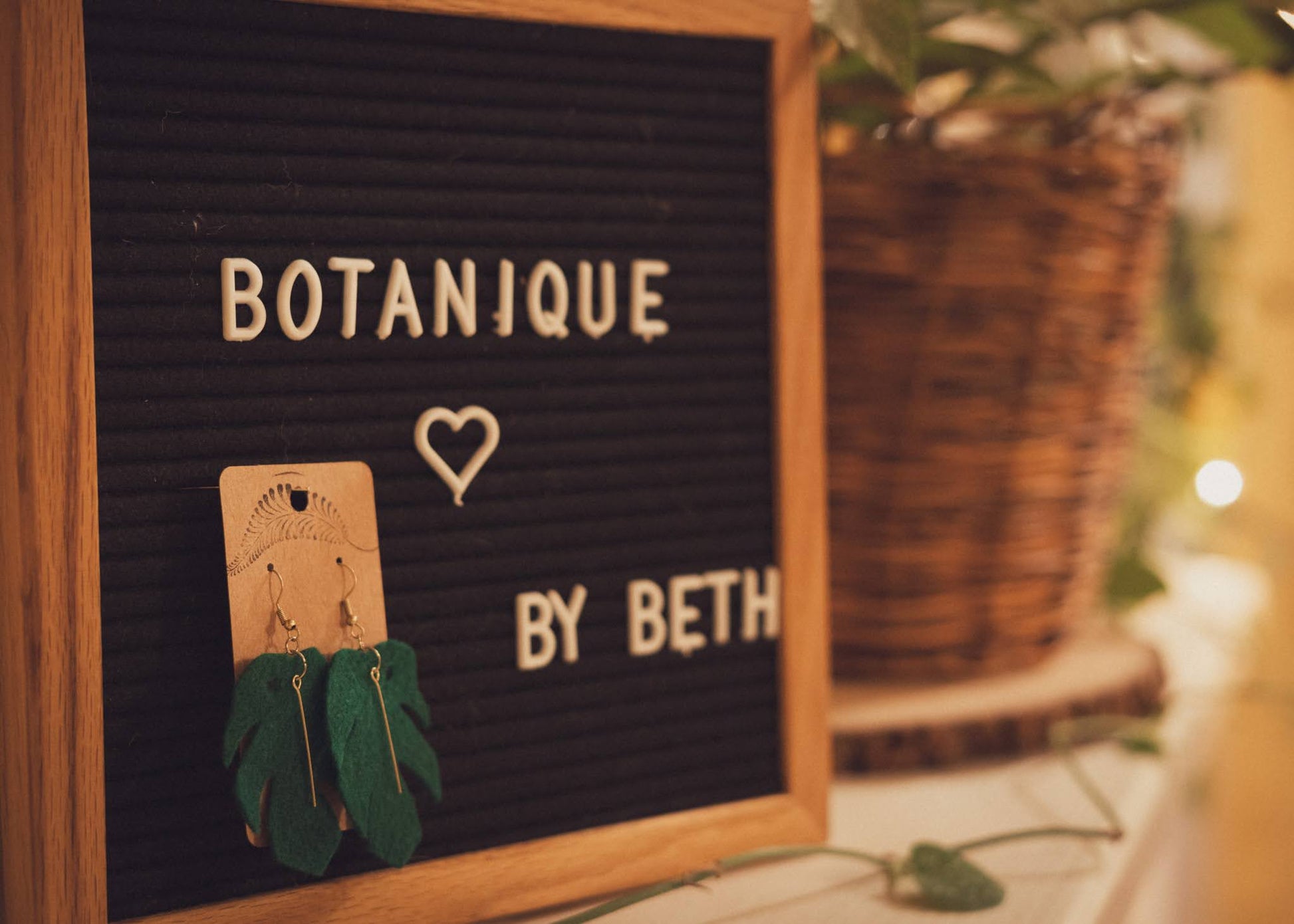 Jungle Monstera leaf felt earrings hanging on a Botanique sign with plants in the background