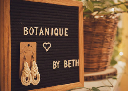 Triple Macrame Loop earrings hanging on a Botanique sign with a gorgeous houseplant on a mantle in the background